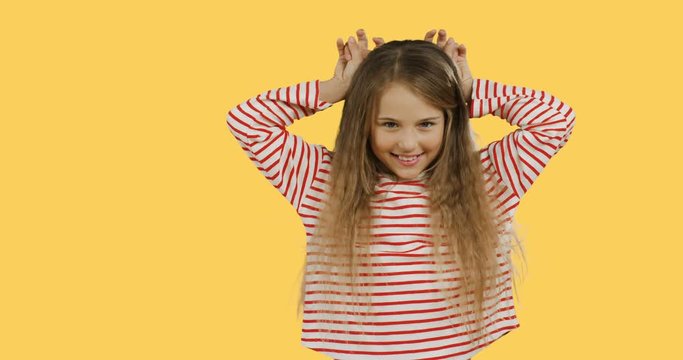Funny blonde small teen girl with long hair standing on the green screen background, having fun and doing rabbit ears gesture. Yellow wall on the back.