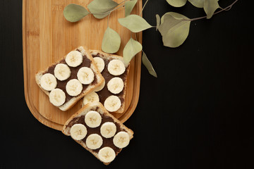 Three banana white bread toasts spread with chocolate butter that lie on a chopping board with a sprig of leaves on a dark background. top view with area for text