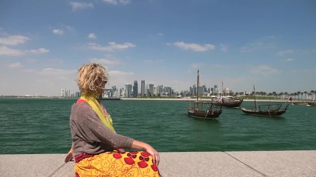 Happy woman on seafront of Doha park along Doha Bay with traditional dhow on background. Lifestyle caucasian tourist enjoys East Mound-Skyline view skyscrapers of Doha Downtown. Qatar, Middle East.