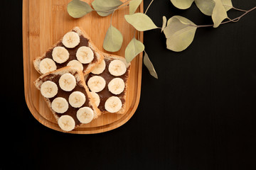 Obraz na płótnie Canvas Three banana white bread toasts spread with chocolate butter that lie on a chopping board with a sprig of leaves on a dark background. top view with area for text