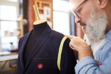 Aged bearded professional tailor using measuring tape while working over new jacket in his workshop