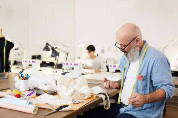 Obraz premium Senior man in shirt and eyeglasses preparing white threads for sewing one of items of new fashion collection while working in tailoring shop