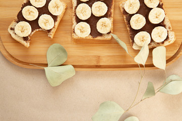 Three banana white bread toasts smeared with chocolate butter that lie on a cutting board with a sprig of leaves on craft paper background. top view with area for text