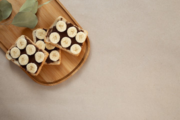 Three banana white bread toasts smeared with chocolate butter that lie on a cutting board with a sprig of leaves on craft paper background. top view with area for text