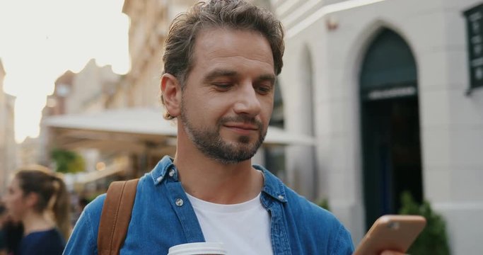 Caucasian Handsome Man With Coffee To Go Walking On The Street And Texting On The Smartphone. Close Up.