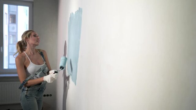 Young girl builder at construction site with paint roller working, painting wall with blue color in apartment