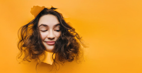 A funny teenage caucasian girl with problematic dry curly hair expresses displeasure on her face. Orange, yellow paper background. Smile, grin.