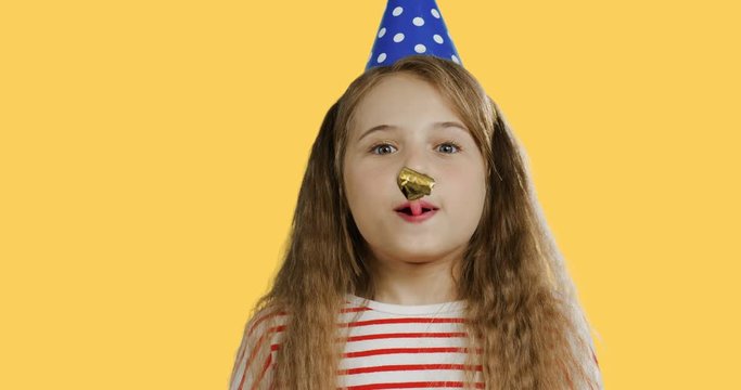 Close up of the funny cute small schoolgirl with a long hair in the birthday party hat and blowing in the horn. Yellow wall back.