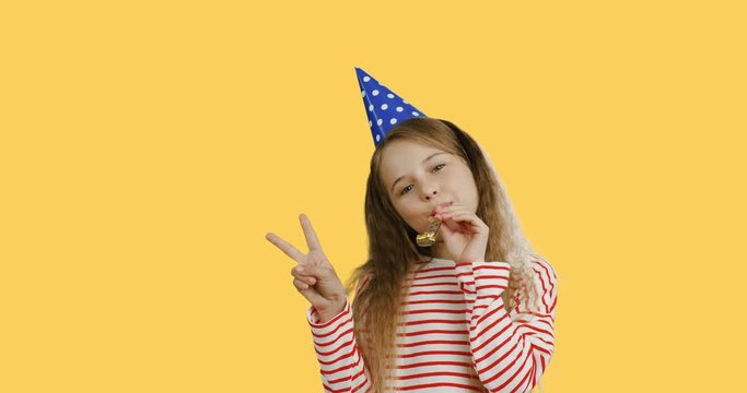 Caucasian cute small teen girl with a long hair in the birthday party hat blowing in the horn and doing victory gesture. Yellow screen background.