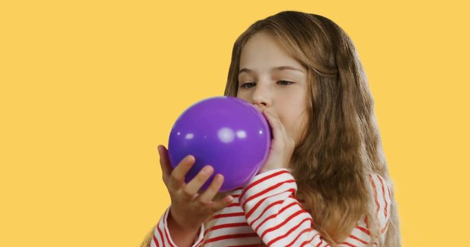 Close up of the blonde pretty small schoolgirl with a long hair inflating a balloon on the yellow screen background.