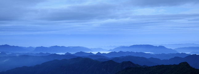 Fototapeta na wymiar Abstract Image, Mountain Silhouettes at dawn - rolling jagged mountain peaks, cold blue color hues. Panoramic Abstract Background Image, overcast skies, layers of rolling mountains in the distance.