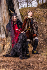 Costume Play Project. Caucasian Couple as Prince and Princess with Black Dog Posing in Medieval Clothing in Spring Forest Outdoors.