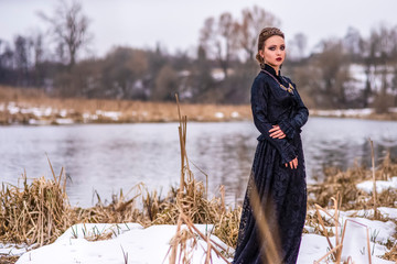 Young Caucasian Brunette Female in Art Dress with Necklace and Crown Posing Against River Outdoors.