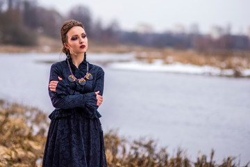 Gorgeous Caucasian Brunette Female in Art Dress with Necklace and Crown Posing Against River...