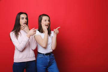 Two lovely young girlfriends dressed in casual clothes looking surprised and showing away with finger isolated over red background.