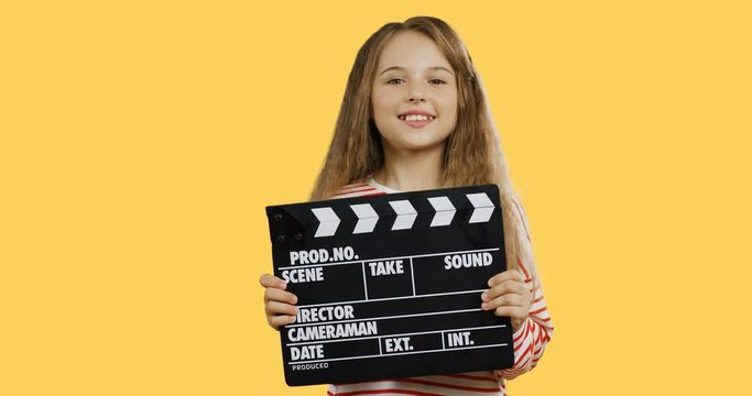 Cute small girl with a long fair hair holding a movie slateboard and clapping it while standing on the yellow screen wall.