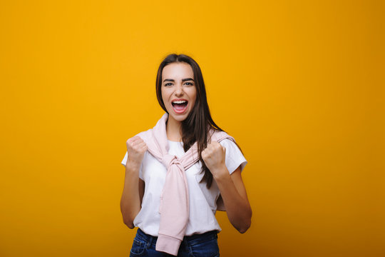 Lovely young cauasian woman with dark long hair looking at camera and screming that she is a winner with clenched fits insolated on a yellow studio wall. Winner concept.