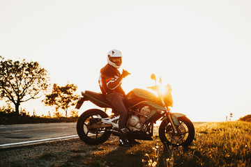 Side view of a young male caucasian biker sitting on the side of the road and looking at the camera and showing that all is ok against sunset while traveling by bike. All is ok concept.