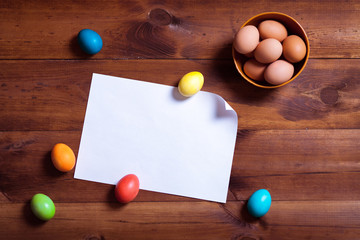 Happy easter background, multicolored eggs and blank white paper on brown wooden table board,...