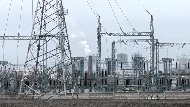 Gray-toned electric substation with a smoking pipe, transformers and power lines against the background of residential high-rise buildings.