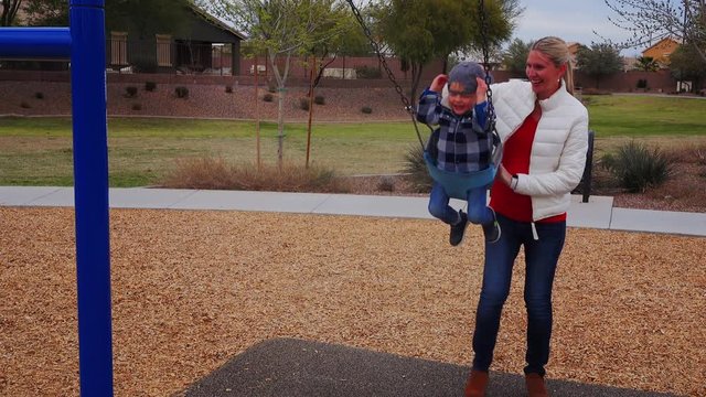A young pregnant mother pushes her son on a swing set in a typical Arizona neighborhood playground on a chilly overcast winter day. Phoenix suburbs.  	