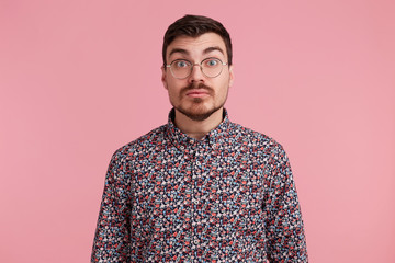 Shocked uncertain surprised man stares through spectacles wearing colorful shirt tries to make situation clear, over pink background