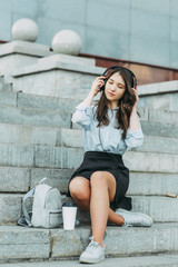 A beautiful student sits on the steps in a shirt and skirt,listening to music in headphones...