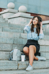 A beautiful student sits on the steps in a shirt and skirt,listening to music in headphones...