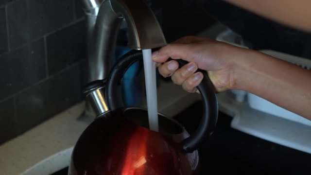 A close up of an African American woman as she fills a used shiny red tea kettle with round black handle and chrome spout with tap water from the faucet in the kitchen sink.