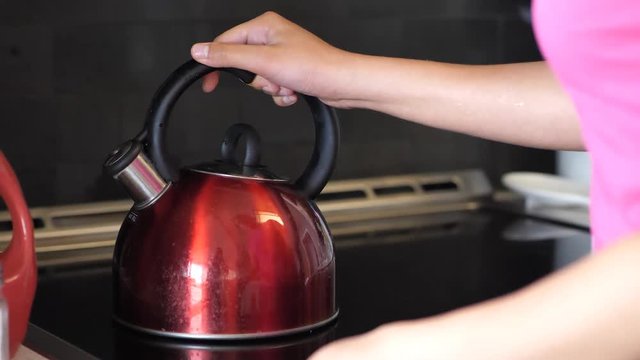 A close up of a mixed ethnicity African American woman wearing a pink shirt as she sets a used shiny red tea kettle with round black handle and chrome spout on a stove to warm up and turns the dial.