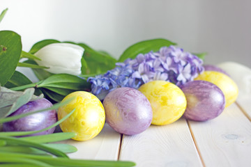 Easter eggs of yellow and lilac color on a wooden background surrounded by greenery, white tulips and hyacinth. Easter background with eggs and tulips. Copy space 