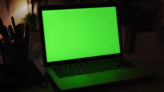 Laptop Computer Showing Green Chroma Key Screen Stands on a Desk in the Living Room. In the Background Cozy Living Room in the Evening with Warm Lights on. Zoom In Shot. 4K