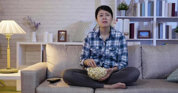 slow movement young asian woman at home sofa couch watching television movie looking sad crying depressed with tv drama film in expressive face gesture. unhappy girl eating popcorn sitting relaxing.