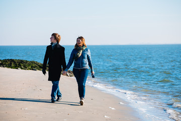 Curly girl and a guy in a coat holding hands. A young couple walks along the sea. Cold season. People are wearing jackets.