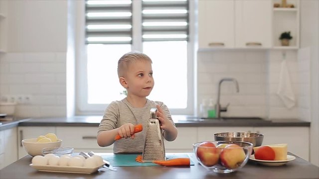 Little beautiful caucasian boy with big blue eyes cooking in the bright kitchen. He is rubs a carrot