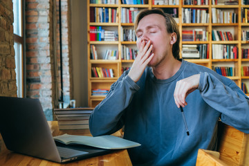 Young student man yawns at work on the computer. The student is tired and wants to sleep while sitting in the library on the background of the bookshelf.