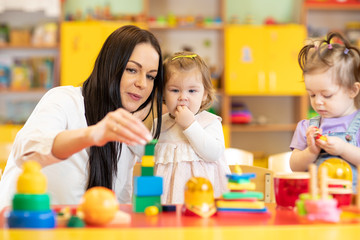 Kindergarten teacher looking after children in daycare. Nursery babies play together with developmental toys.