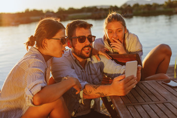 Two young woman and man using smartphone by the river and smiling
