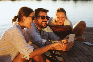 Two young woman and man using smartphone by the river and smiling