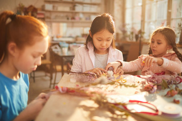Multi-ethnic group of little girls creating spring wraths and decorations made of flowers in art class, scene lit by serene sunlight