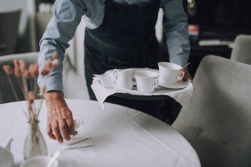 Fototapeta premium Waiter in apron holding tray with cups of coffee and placing sugar on table