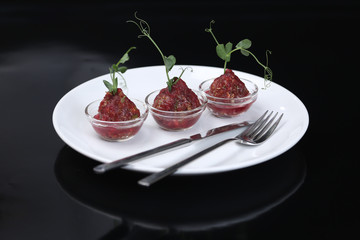 Red appetizing Tartar sauce in three pialas on a plate with a knife and fork, decorated with a stem of sprouted green peas, close-up, for a menu