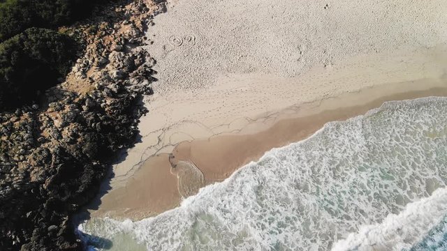 an aerial shot looking down on blue water waves splashing onto the shore of a sandy beach. Cala Varques, Majorca, Spain