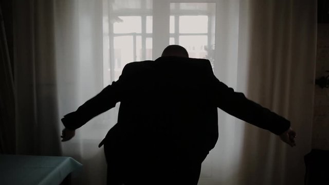 Man puts on black jacket standing before window.