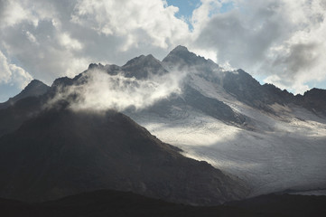 Mountain landscape of a high mountain slope with a cracked glacier muddy volcanic slope against the backdrop of the Caucasus Mountains in the window of clouds. Glaciers of the North Caucasus