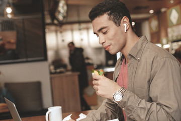 Handsome afro american guy eating sandwich and using wireless earphones