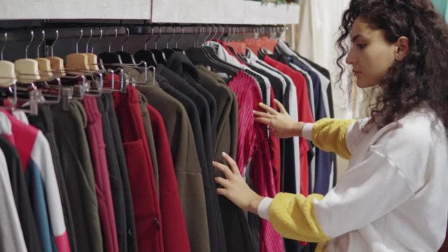 Woman is viewing clothing on sale in shop, moving hangers with pants and sweater