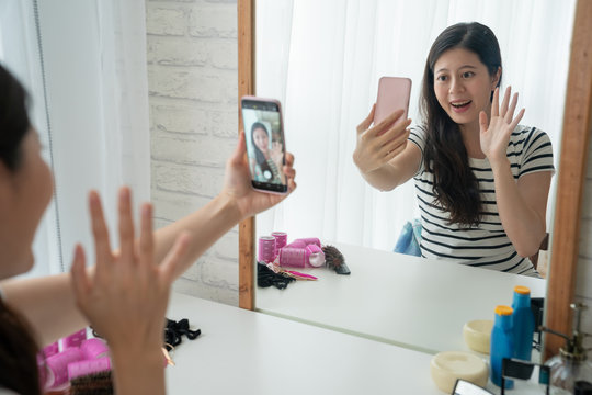 Beauty asian blogger filming makeup tutorial with smartphone in front of mirror at dressing table indoors. young woman waving hands starting recording video get ready in the morning bright bedroom.