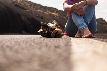 Ground alternative point of view of woman sitting on the road with nude feet and a broken pair of shoes near her - concept of walking for alternative lifestyle vacation - travel and enjoy mountains 