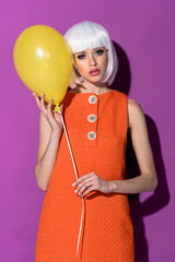 Stylish young woman in white wig holding yellow air balloon on purple background